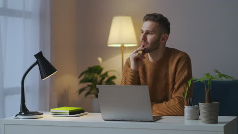 young-guy-is-wearing-brown-sweater-is-sitting-in-front-of-table-with-laptop-at-evening-working-at-home-creative-profession-and-remote-work